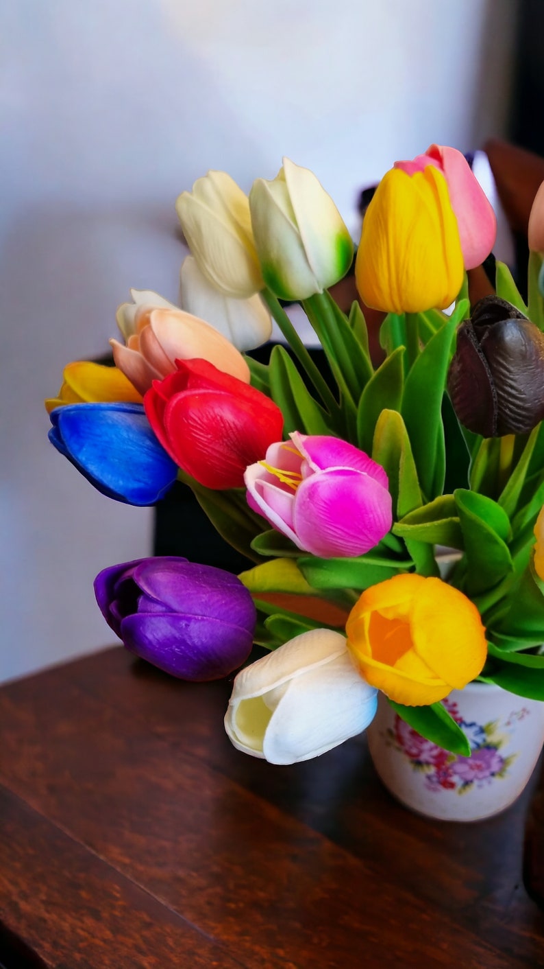 May include: A bouquet of colorful artificial tulips in a white ceramic vase with a floral pattern. The tulips are in shades of red, pink, yellow, blue, purple, and white.