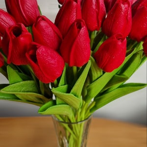 Peut inclure: Un bouquet de tulipes rouges dans un vase en verre transparent. Les tulipes sont dispos&eacute;es en &eacute;ventail, les tiges pointant vers le bas. Le vase est pos&eacute; sur une table en bois.