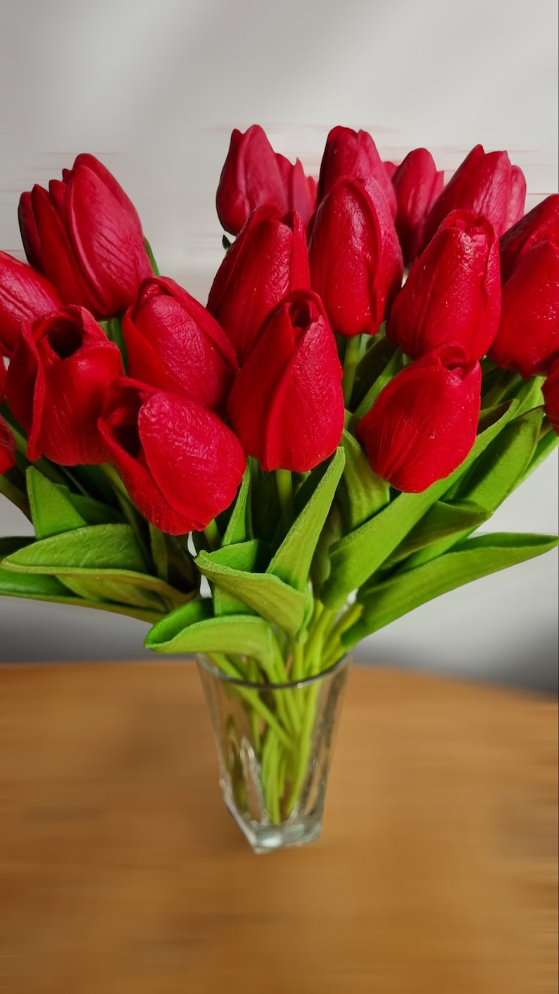 Peut inclure: Un bouquet de tulipes rouges dans un vase en verre transparent. Les tulipes sont dispos&eacute;es en &eacute;ventail, les tiges pointant vers le bas. Le vase est pos&eacute; sur une table en bois.