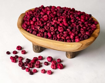 May include: A wooden bowl with a natural light brown finish and three short, thick legs, filled with a large quantity of small, dried, deep magenta-colored pumpkin-shaped decorative items, with several more scattered on the surface below.