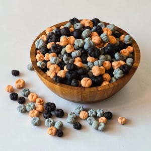 Könnte beinhalten: Eine große Holzschale gefüllt mit einer Sammlung kleiner, kürbisförmiger Dekorationen in den Farben Orange, Schwarz und gedämpftem Blaugrau. Mehrere zusätzliche Miniatur-Kürbisdekorationen sind außerhalb der Schale verstreut.
