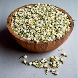 May include: A wooden bowl filled with dried jasmine flowers. The flowers are white with green stems and buds. Some flowers are scattered on the grey surface. The bowl is made of light brown wood.