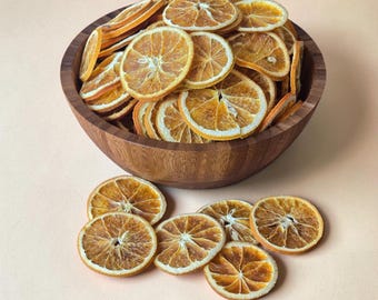May include: A dark brown wooden bowl filled with numerous thin, dehydrated orange slices. Several additional dried orange slices are scattered on the surface in front of the bowl, showcasing their circular shape, textured pulp, and vibrant orange rinds.