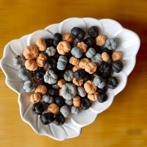 May include: A white ceramic bowl filled with small, decorative pumpkins in shades of orange, gray, and black. The bowl is on a wooden surface.