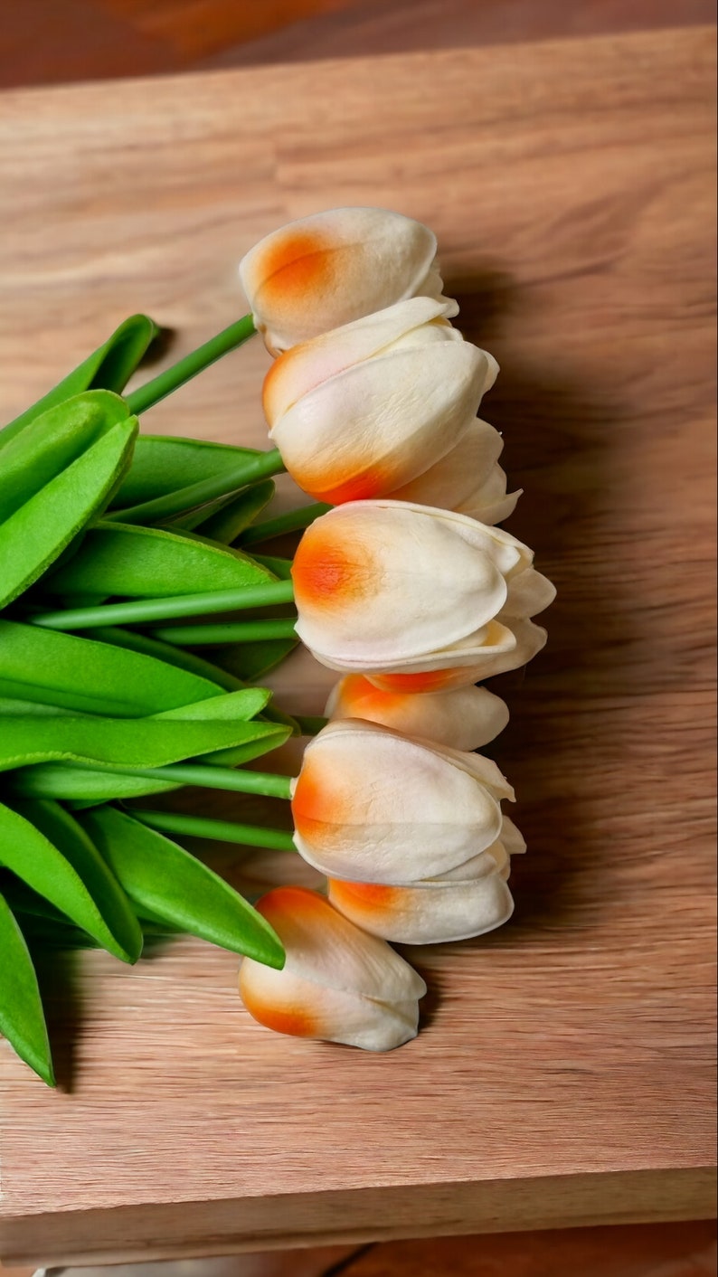 Peut inclure: Un bouquet de tulipes blanches avec des pointes orange, dispos&eacute; sur une surface en bois. Les tulipes ont des tiges et des feuilles vertes.