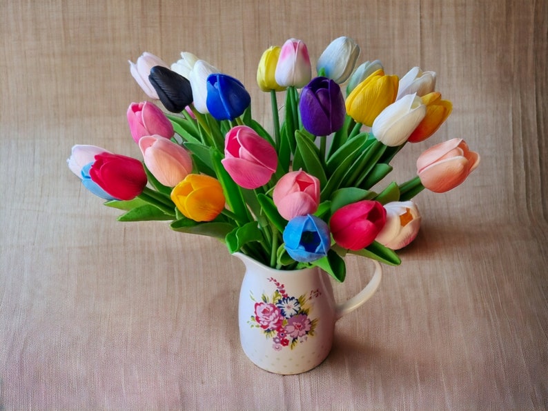 Puede incluir: Un ramo de tulipanes artificiales de colores en una jarra blanca con un patr&oacute;n floral. Los tulipanes son en tonos de rosa, morado, azul, amarillo, blanco y naranja.