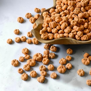 Puede incluir: Una gran colección de pequeñas cuentas de color naranja con forma de calabaza hechas de un material mate. Muchas de estas cuentas están amontonadas dentro de un cuenco de madera de color marrón claro, mientras otras están esparcidas.