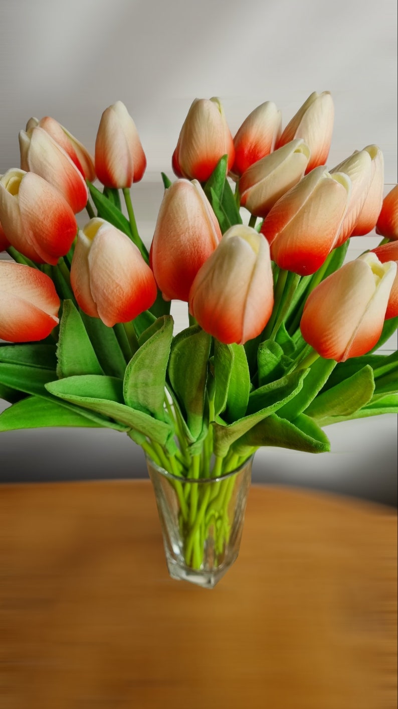 Peut inclure: Un bouquet de tulipes artificielles dans un vase en verre transparent. Les tulipes sont un m&eacute;lange de blanc et d'orange avec un effet d&eacute;grad&eacute;. Les tiges sont vertes.