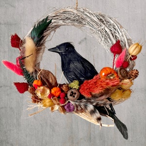 Peut inclure: Une couronne de brindilles blanches d&eacute;cor&eacute;e de feuillage d'automne, y compris des baies rouges, des citrouilles orange et des pommes de pin brunes. Un corbeau noir est perch&eacute; sur la couronne. La couronne est suspendue &agrave; une ficelle. Le texte "Fluff & Co" est visible en bas de l'image.