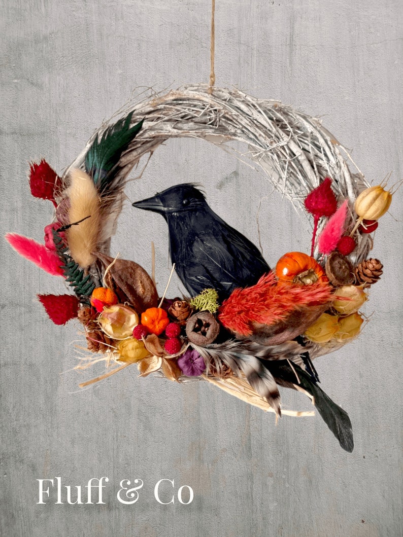 Peut inclure: Une couronne de brindilles blanches d&eacute;cor&eacute;e de feuillage d'automne, y compris des baies rouges, des citrouilles orange et des pommes de pin brunes. Un corbeau noir est perch&eacute; sur la couronne. La couronne est suspendue &agrave; une ficelle. Le texte "Fluff & Co" est visible en bas de l'image.
