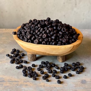 May include: A light brown wooden bowl with three short legs, filled with a large quantity of small, matte black pumpkin-shaped decorative items. Several additional black pumpkin-shaped pieces are scattered around the base of the bowl.