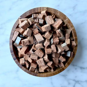 Peut inclure: Un bol en bois rempli de morceaux de coque de noix de coco bruns. Les morceaux de coque de noix de coco sont coupés en petites formes irrégulières. Le bol est en bois clair et repose sur une surface de marbre blanc et gris.