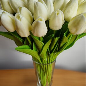Peut inclure: Un bouquet de tulipes artificielles blanches dans un vase en verre transparent. Les tulipes sont dispos&eacute;es dans un vase sur une surface en bois.