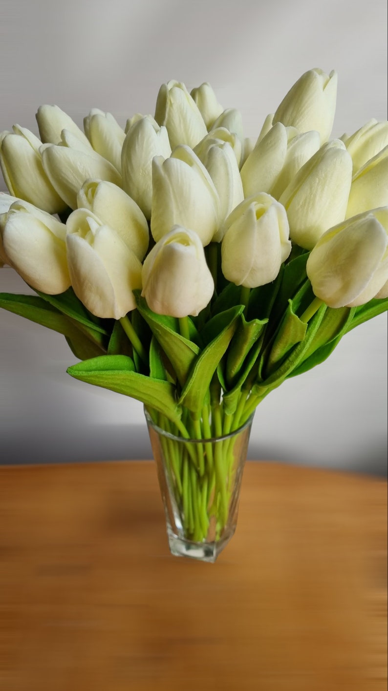 Peut inclure: Un bouquet de tulipes artificielles blanches dans un vase en verre transparent. Les tulipes sont dispos&eacute;es dans un vase sur une surface en bois.