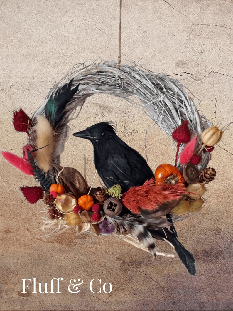 Peut inclure: Une couronne de brindilles d&eacute;cor&eacute;e de feuillage d'automne, y compris des baies rouges, des citrouilles orange et des fleurs s&eacute;ch&eacute;es, avec un corbeau noir perch&eacute; dessus. La couronne est suspendue &agrave; une ficelle et le texte "Fluff & Co" est visible en bas de l'image.