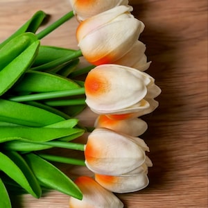 May include: A bouquet of white tulips with orange tips, arranged on a wooden surface. The tulips have green stems and leaves.