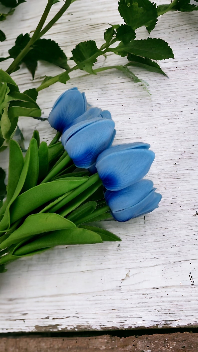 Puede incluir: Un ramo de tulipanes azules con tallos y hojas verdes sobre un fondo de madera blanca.