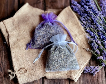 May include: Two small lavender-filled sachets, one purple and one white, are on a burlap surface with a bunch of lavender flowers to the right.