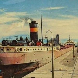 May include: Vintage postcard featuring the freighter "Frank Purnell" of Wilmington, passing through a canal. The ship is brown and white with a black and orange smokestack. The sky is blue with clouds.