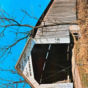 May include: A white wooden covered bridge with a sign that reads "Cross This Bridge At A Walk" and the number "1914". The bridge is set against a bright blue sky.