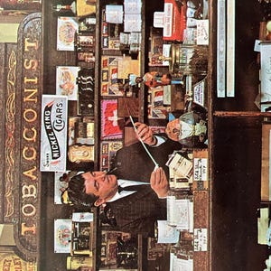 May include: A vintage tobacco shop interior with a wooden counter and shelves stocked with various tobacco products. A man in a suit is standing behind the counter, holding a long, thin object. The shop sign reads "TOBACCONIST" and "SMOKE NICKEL KING CIGARS".