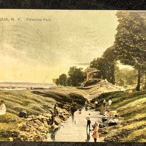 May include: Vintage postcard featuring a scenic view of Chautauqua, N.Y., Palestine Park. The image shows a stream flowing through a grassy area, with people enjoying the outdoors. A building sits atop a small hill in the background.