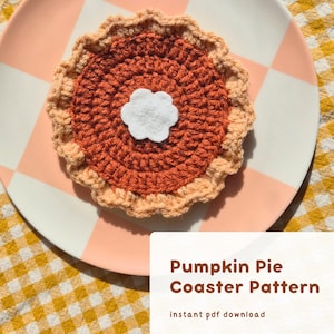 May include: A crocheted pumpkin pie coaster with a white felt dollop of whipped cream, resting on a pink and white checkered plate. The coaster is orange and tan, with the text "Pumpkin Pie Coaster Pattern" and "instant pdf download".