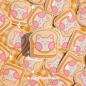 May include: A selection of stickers depicting a cartoon cat face on a slice of toast. The cat has pink cheeks and a closed-mouth smile. The toast is light brown with a white cat face and pink accents. The stickers are scattered on a yellow and white checkered surface.