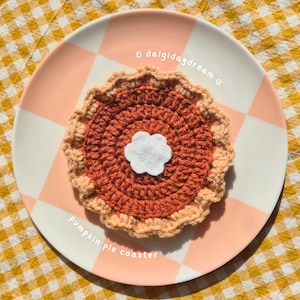 May include: A crocheted pumpkin pie coaster with a white felt cloud in the centre, resting on a pink and white chequered plate. The text "Pumpkin pie coaster" is visible. The coaster is handmade.