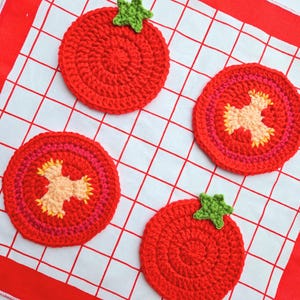 Peut inclure: Quatre dessous de verre en crochet en forme de tomates. Les dessous de verre sont rouges avec des dessus verts, et certains ont un design en coupe transversale avec des détails roses, jaunes et pêche. Ils reposent sur un tissu à carreaux blanc et rouge.