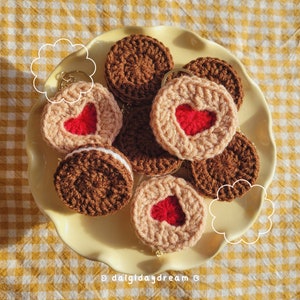 May include: A collection of crocheted cookie keychains in shades of brown and beige, some with red heart cutouts. One keychain has a white filling. The keychains are arranged on a yellow plate, with a yellow and white checkered background.