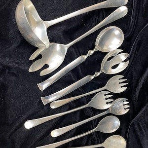 May include: A set of eight silver serving utensils, including a ladle, two serving spoons, and five serving forks. The utensils are arranged on a black fabric background.