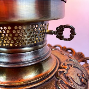 May include: Close-up of a brass lamp base with a keyhole and a decorative, ornate design. The base has a round, perforated top and a detailed, intricate bottom.