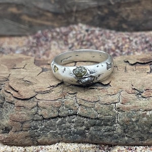 Op de afbeelding: Een zilveren ring met een gehamerde afwerking en groene en gouden edelstenen die in de band zijn gezet.