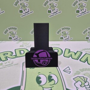 May include: A black phone stand with a purple logo that says "lenscollectables". The stand is on a green surface with a cartoon character on it.