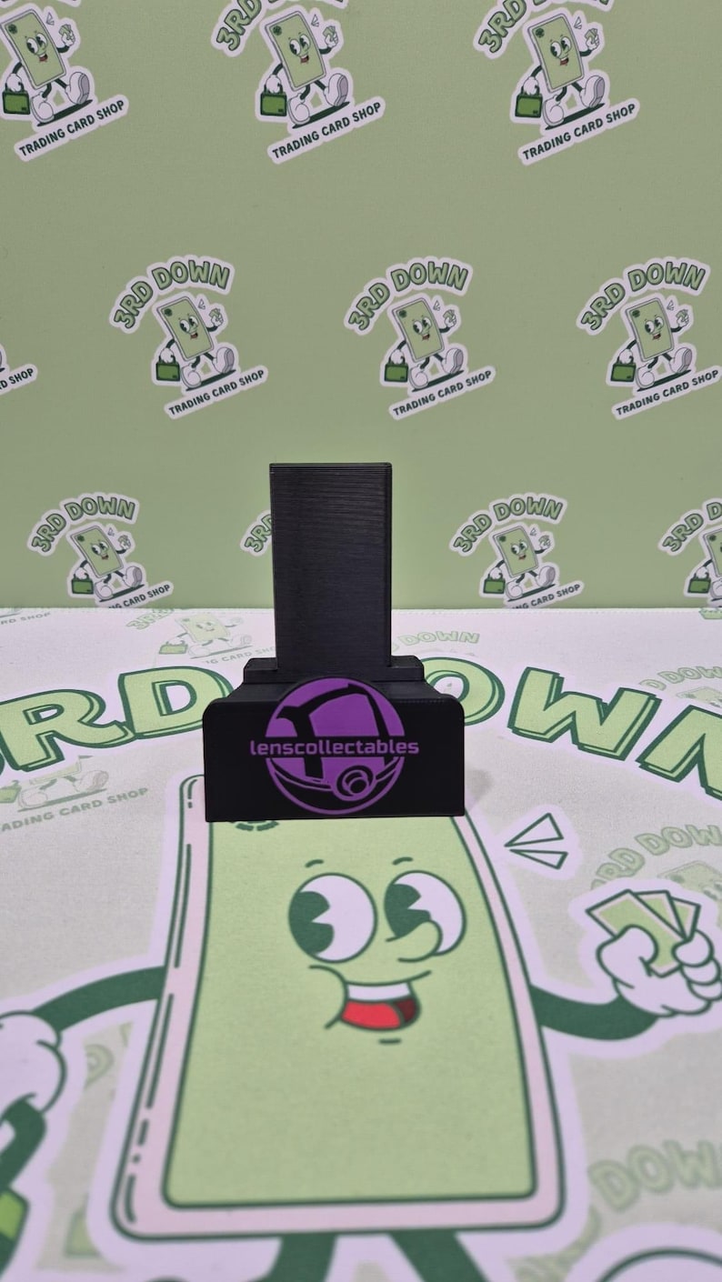 May include: A black phone stand with a purple logo that says "lenscollectables". The stand is on a green surface with a cartoon character on it.
