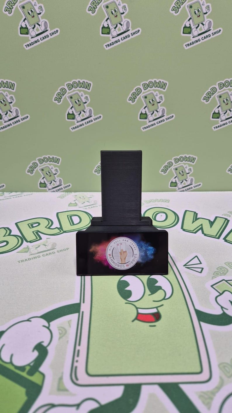 May include: A black and white card stand with a colorful background featuring the text "Hammonds Hobbies". The stand is on a green surface with multiple stickers that say "3rd Down Trading Card Shop".