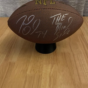 Puede incluir: Un balón de fútbol americano de cuero marrón con el logotipo de la NFL y las firmas de "Roy" y "The Game" en tinta blanca.