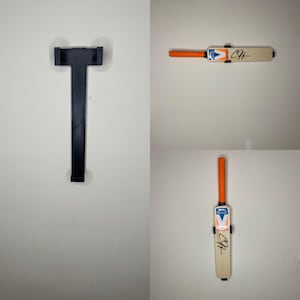 Puede incluir: Un soporte de pared negro para un bate de críquet y dos bates de críquet. Los bates tienen mangos naranjas, logotipos blancos y azules y una firma. Los bates de críquet están montados en una pared blanca.