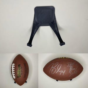 Könnte beinhalten: Ein brauner Lederfußball mit weißen Nähten und dem aufgedruckten Text "Wilson" und "NFL". Der Fußball ist mit der Nummer "79" und dem Text "THE" und "MIN" in schwarzem Stift signiert. Der Fußball wird auf einem schwarzen Plastikständer präsentiert.