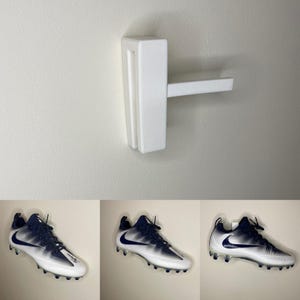 Puede incluir: Un soporte de pared de plástico blanco para zapatos con una zapatilla de fútbol blanca colgada. La zapatilla es blanca con detalles negros y azules.