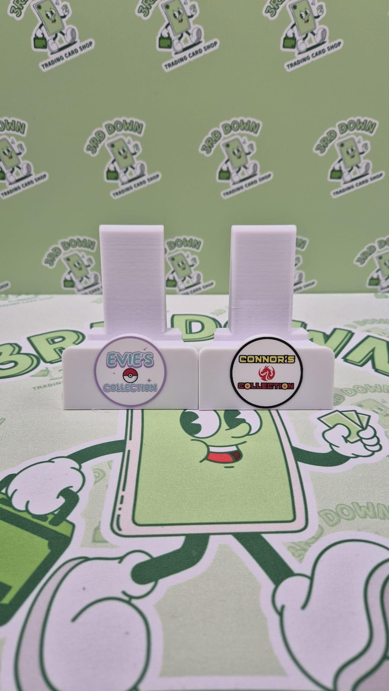 May include: Two white card holders with a round sticker on the front. The left sticker is purple with the text "Evie's Collection" and a Pokemon character. The right sticker is yellow with the text "Connor's Collection" and a fire symbol.