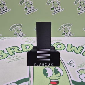 May include: A black card holder with a white stripe design and the text "SLABZUK" on the front. The holder is on a green background with a repeating pattern of a cartoon character.