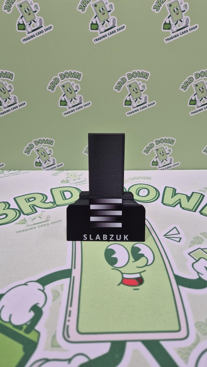 May include: A black card holder with a white stripe design and the text "SLABZUK" on the front. The holder is on a green background with a repeating pattern of a cartoon character.