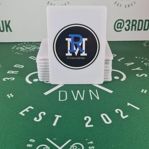 Könnte beinhalten: Ein Stapel weißer quadratischer Karten mit einem schwarzen und blauen Logo, das auf der obersten Karte "RM Collectables" lautet. Das Logo ist ein Kreis mit einem blauen "R" und einem weißen "M" darin. Die Karten liegen auf einer grünen Oberfläche mit einem weißen Logo, das "3RD DOWN EST 2021" lautet.