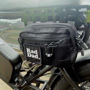 May include: Black utility bag attached to a bicycle. The bag has a zippered main compartment, a front patch with the text "Rad Dad," and a hook-and-loop closure. The bag is mounted on a black metal rack.