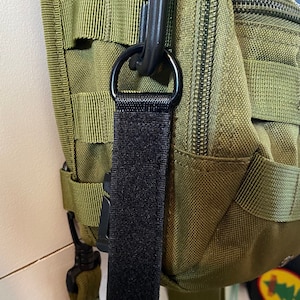 May include: A black nylon strap with a metal D-ring attached to a green tactical backpack. The strap is secured with a black hook and loop fastener.