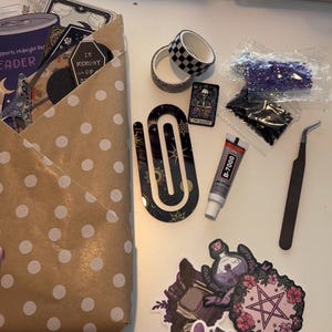 May include: A craft kit containing a brown polka dot paper envelope, two rolls of patterned washi tape, a large black U-shaped acrylic bookmark, a tube of B-7000 glue, metal tweezers, two small bags of beads, and several stickers featuring occult imagery.