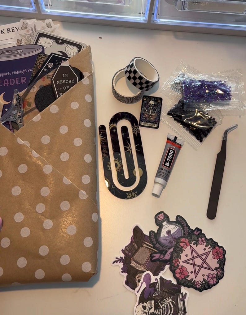 May include: A craft kit containing a brown polka dot paper envelope, two rolls of patterned washi tape, a large black U-shaped acrylic bookmark, a tube of B-7000 glue, metal tweezers, two small bags of beads, and several stickers featuring occult imagery.