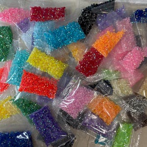 May include: A collection of small, clear plastic bags filled with multi-colored, faceted resin rhinestones. The assortment includes vibrant shades of red, orange, yellow, green, blue, purple, pink, black, and silver, all featuring a reflective finish.